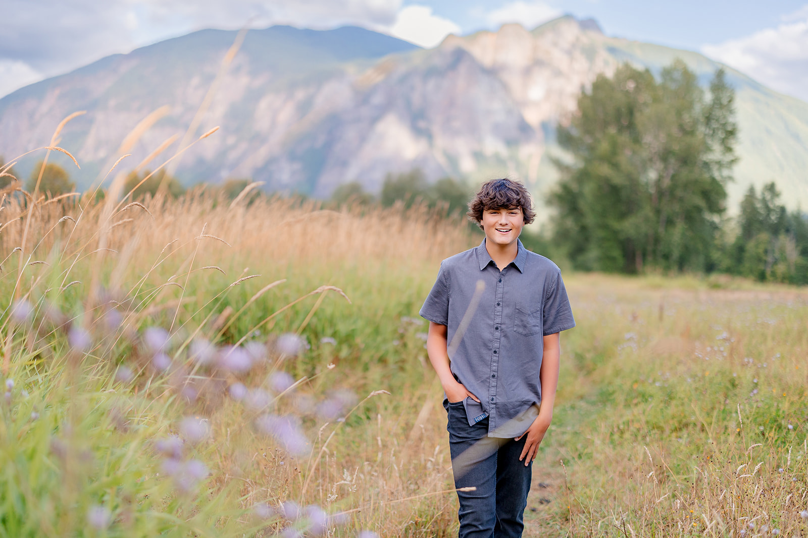 A happy boy in a grey shirt and jeans walks on an overgrown trail by the mountains