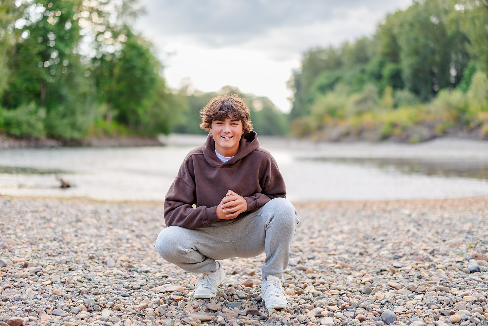 A high school senior squats on a rocky lakeshore in a brown hoodie and grey pants after some SAT Prep in Kirkland