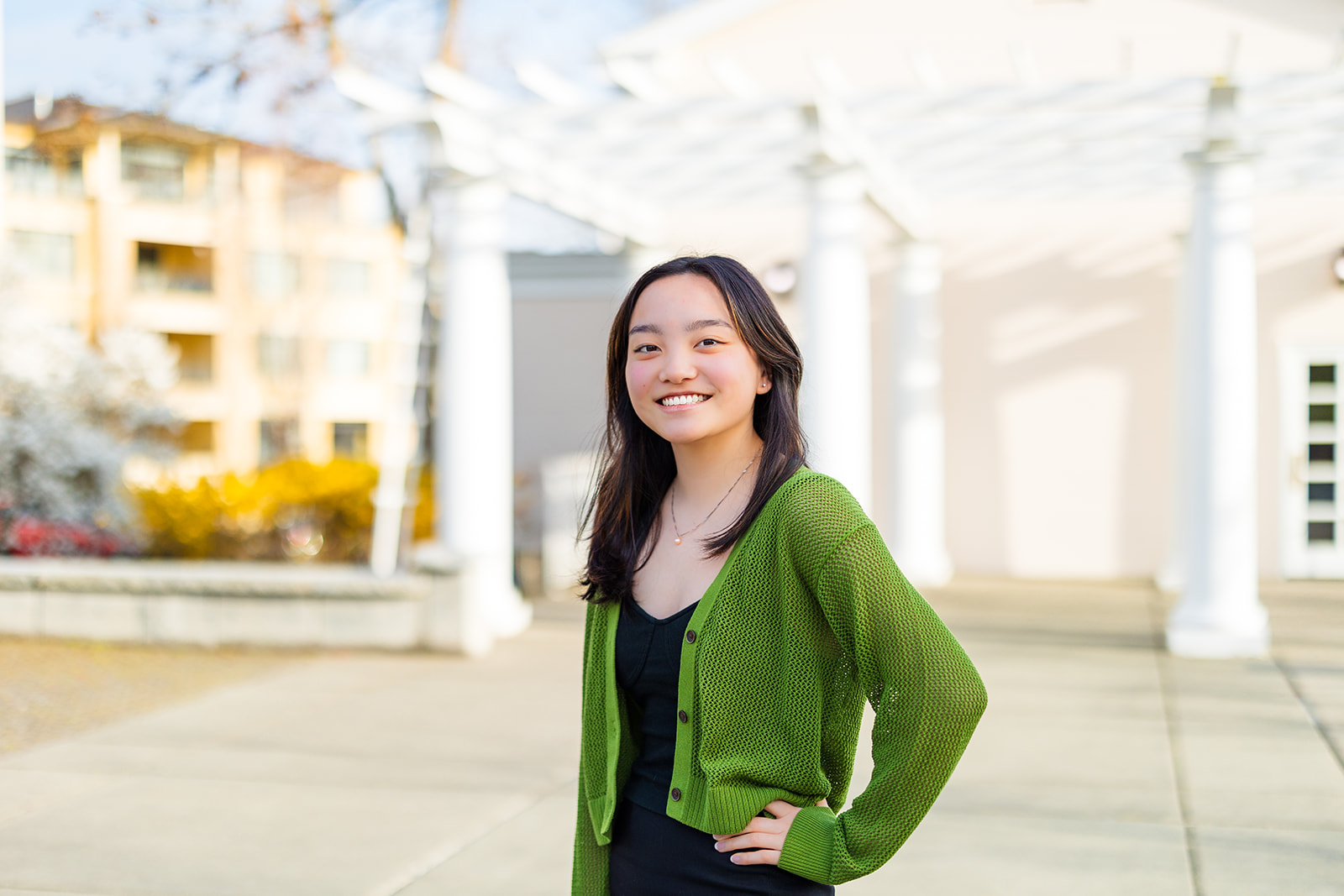 A happy high school senior stands in a black dress and green sweater on a patio with a hand on her hip