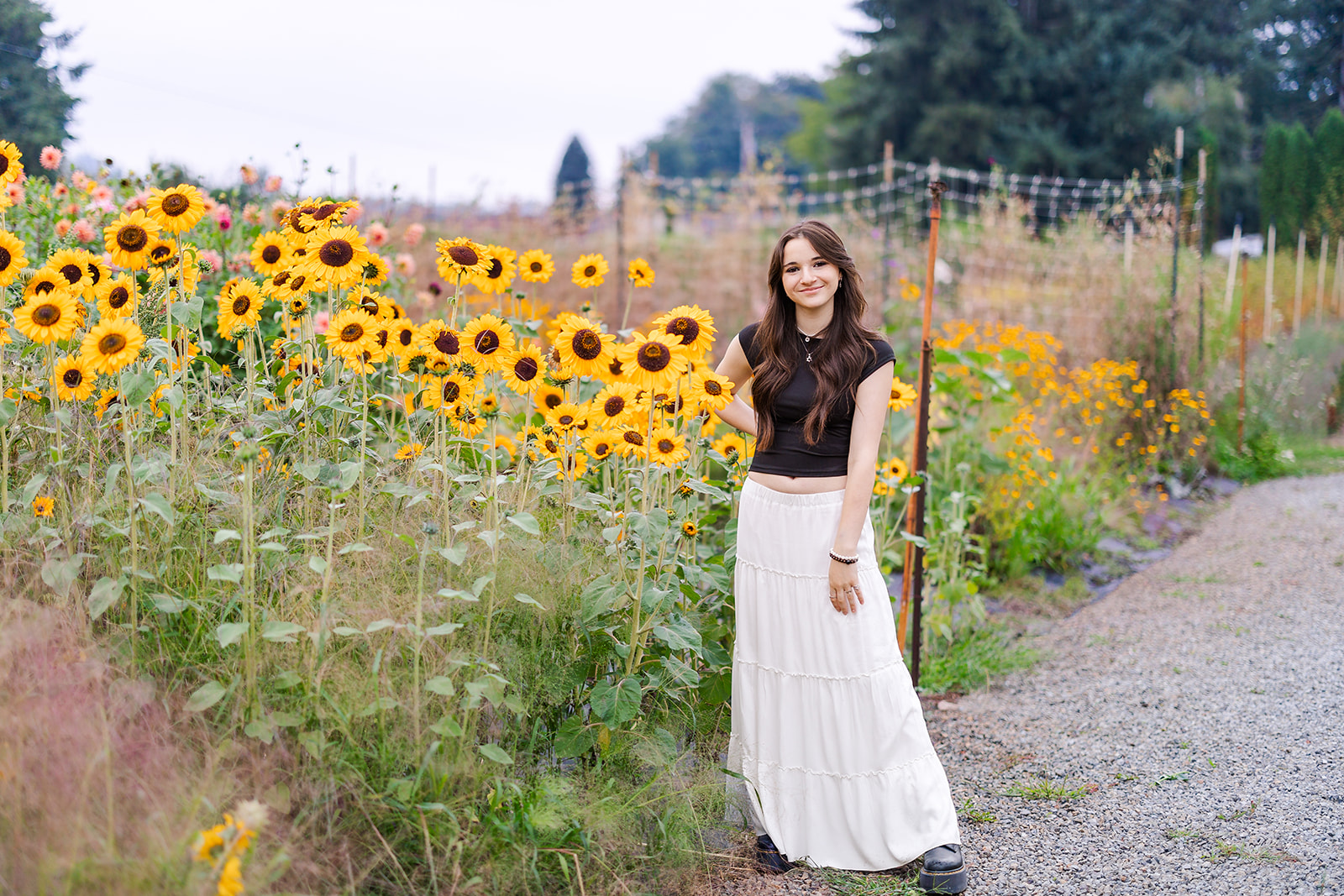 A high school senior in a black shirt and long white skirt stands by some sunflowers in a garden after visiting Seattle Hair Salons