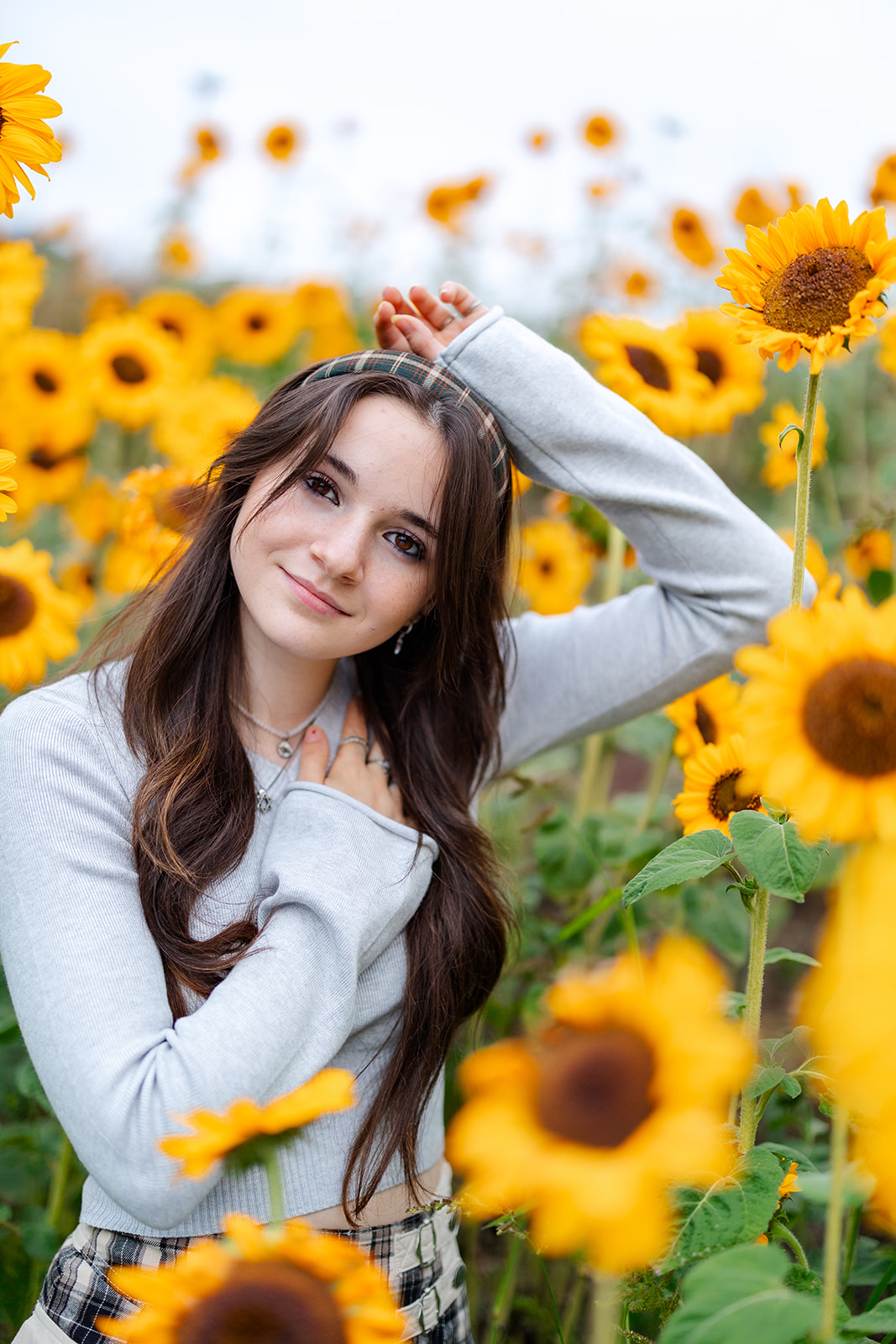 A high school senior with brown hair stands in a field of sunflowers in a grey sweater after visiting Seattle Hair Salons