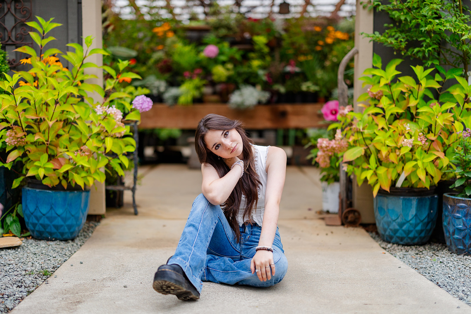 A teenage girl in jeans and boots sits at the entrance to a greenhouse