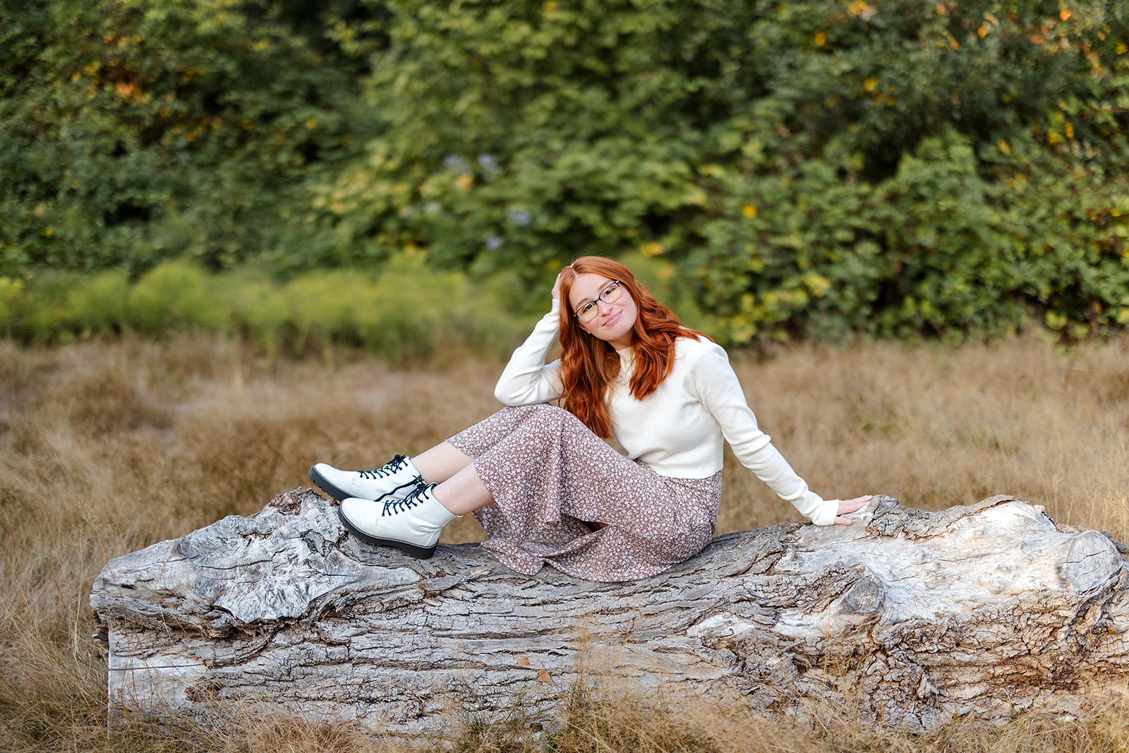 A red hair high school senior sits on a large log in a dress and white sweater