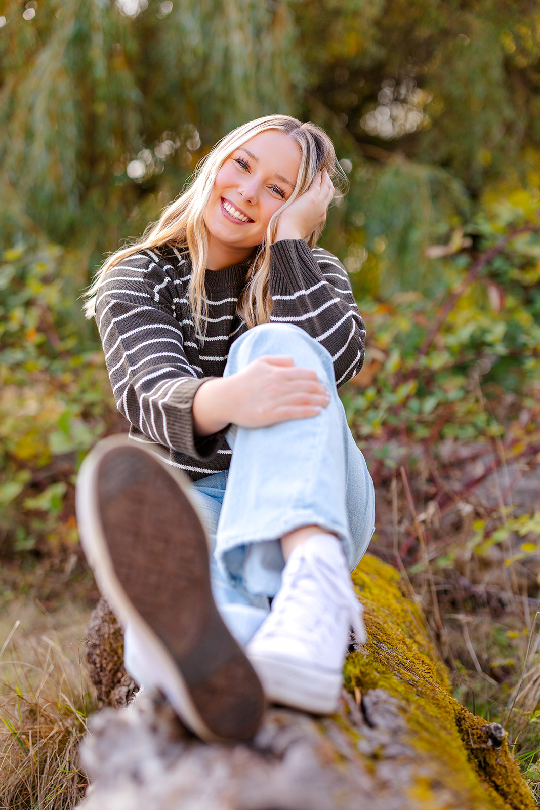 A high school senior in jeans and a stripe knit sweater sits along a log smiling after attending Seattle Private Schools