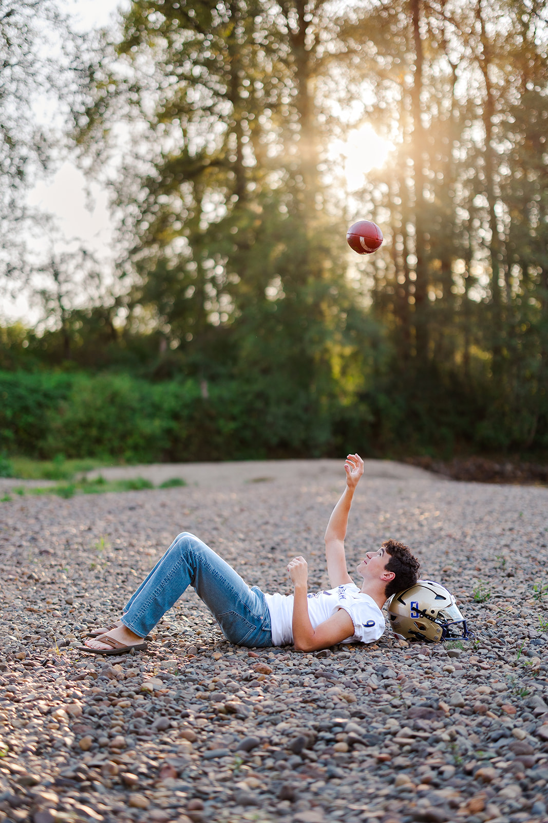 A high school senior lays on pebbles by a river tossing a football over his head at sunset