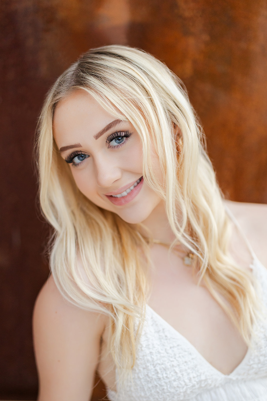 A blonde high school senior smiles while sitting against a rusted wall in a white top before visiting Tanning Salons Renton, WA