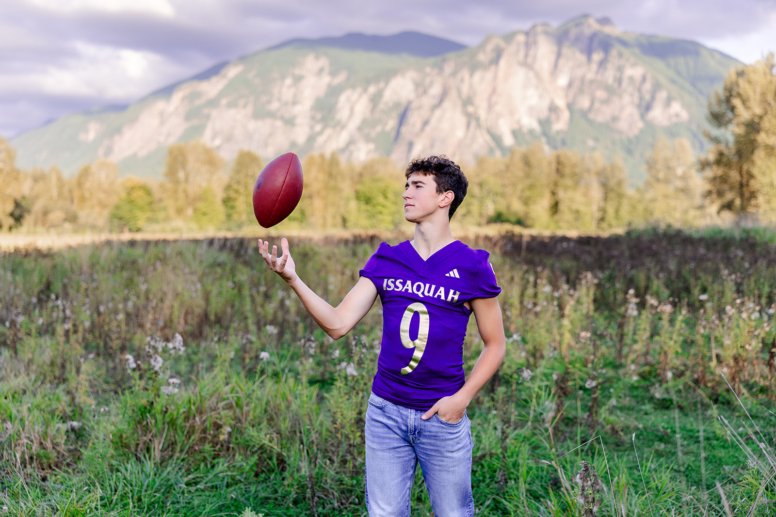 A high school senior in a football jersey tosses a football while standing in a remote field by the mountains after finding Tutoring Issaquah