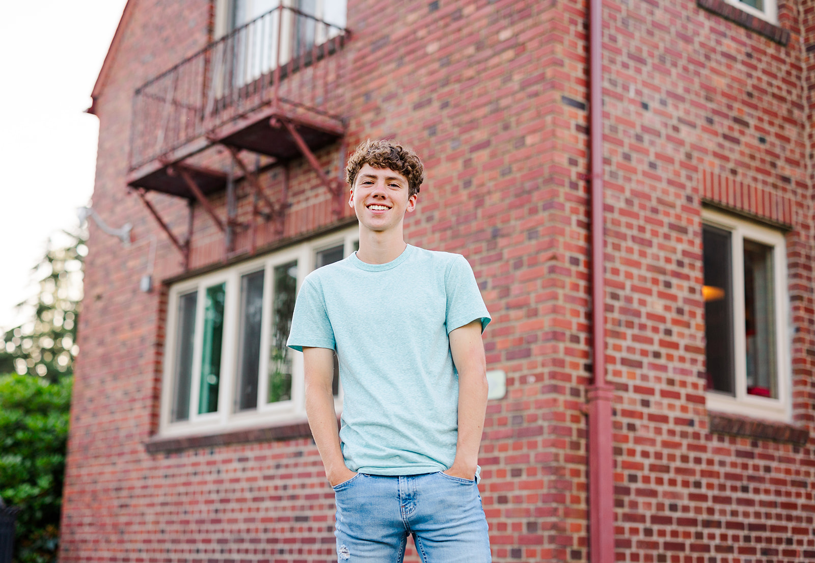 A happy man in jeans and green shirt stands by a brick building with hands in pockets