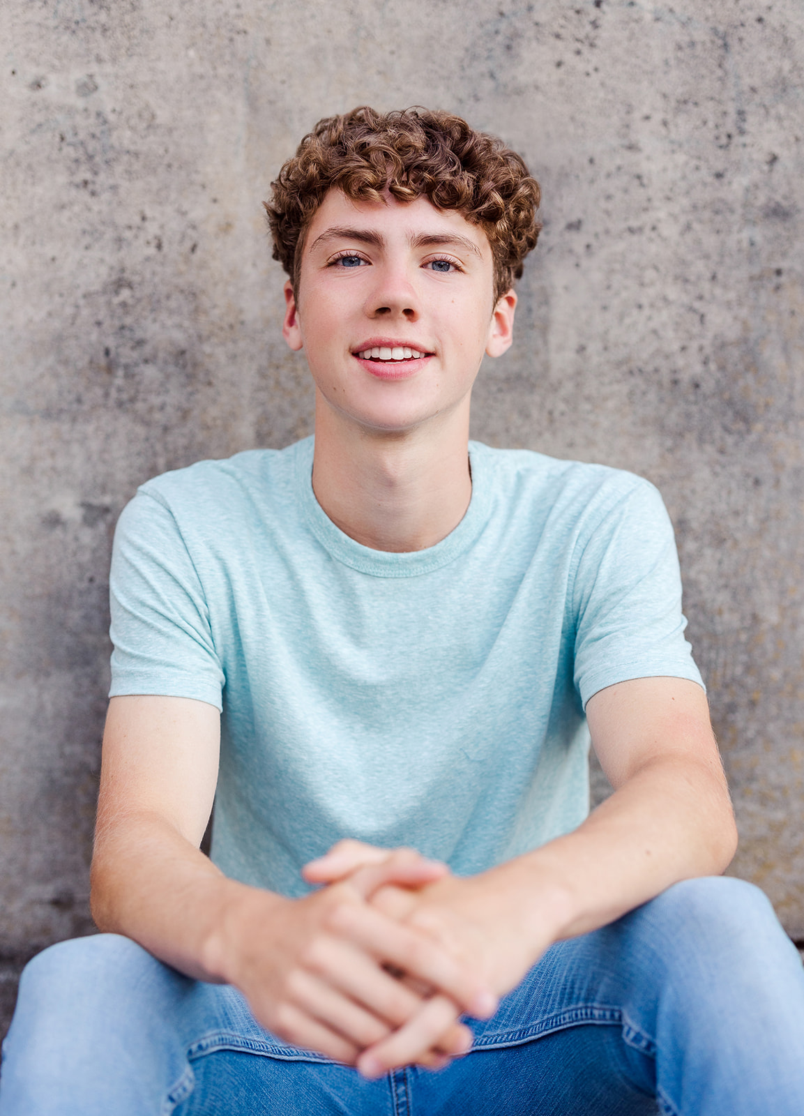 A smiling high school senior boy sits against a wall in a blue shirt and jeans after some Tutoring Renton, WA