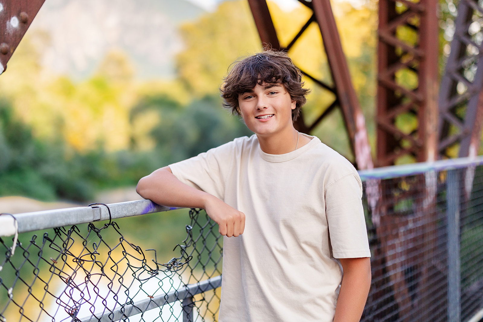 A smiling high school senior in a beige shirt leans on a fence on a steel bridge over a river after finding Tutoring Seattle