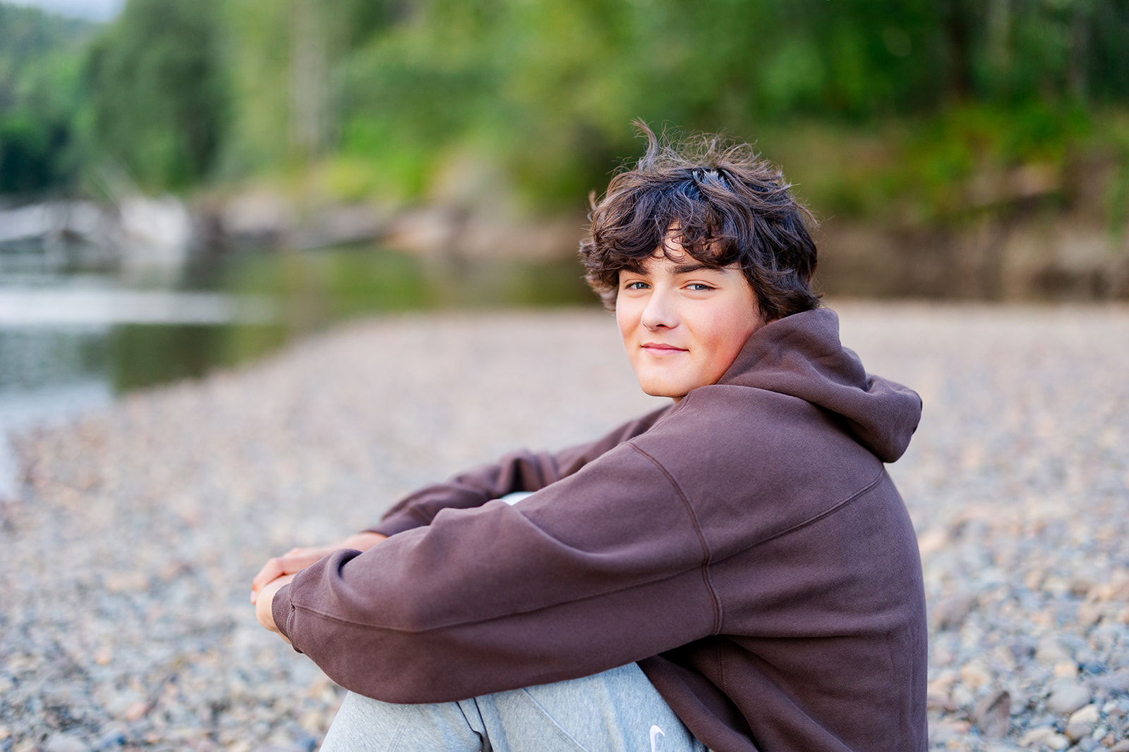 A high school senior in a brown hoodie sits on a rocky beach at sunset after getting Tutoring in Seattle