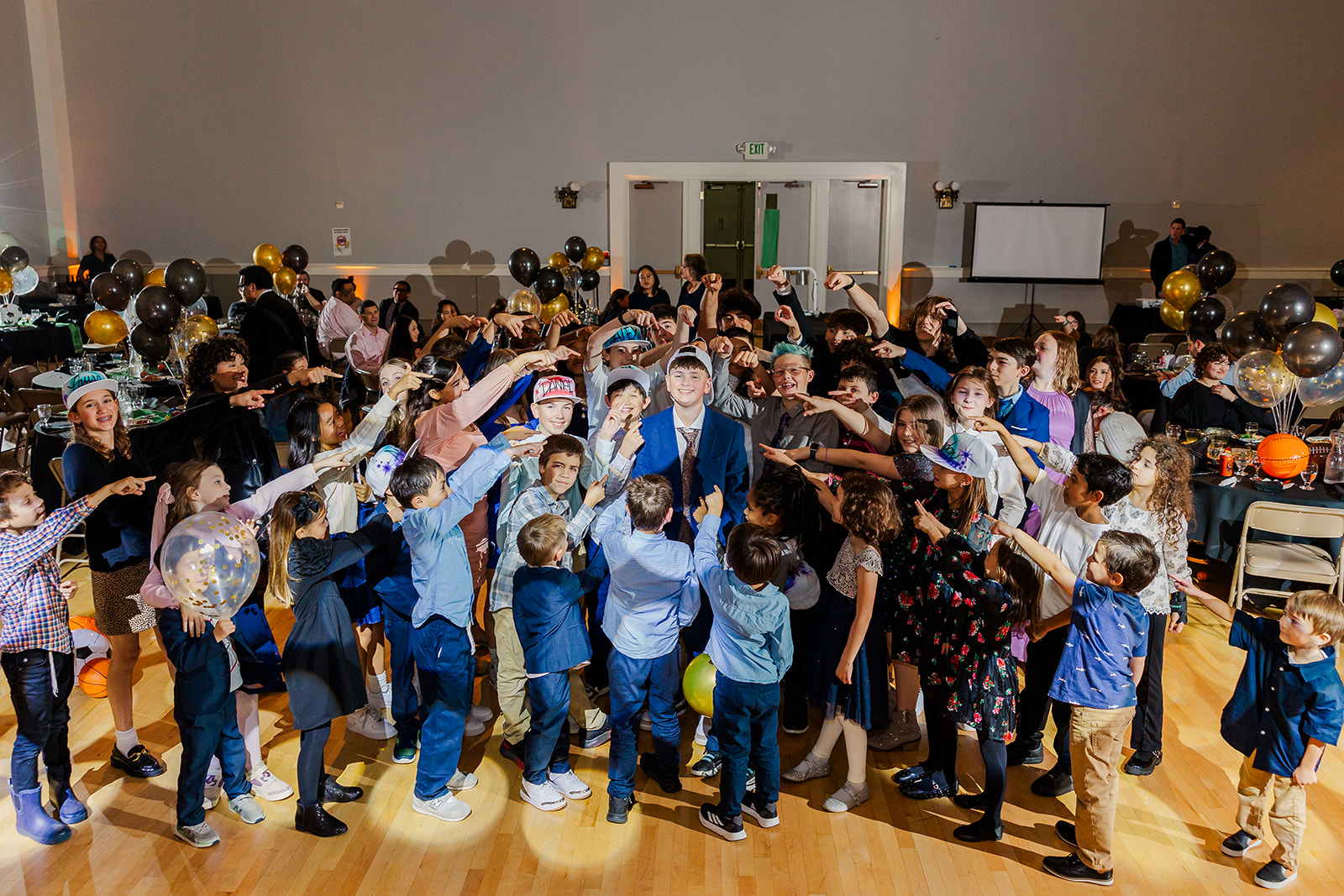 A crowd of young kids point to a smiling boy in the center in a blue suit and backwards hat planned by Pink Blossom Events