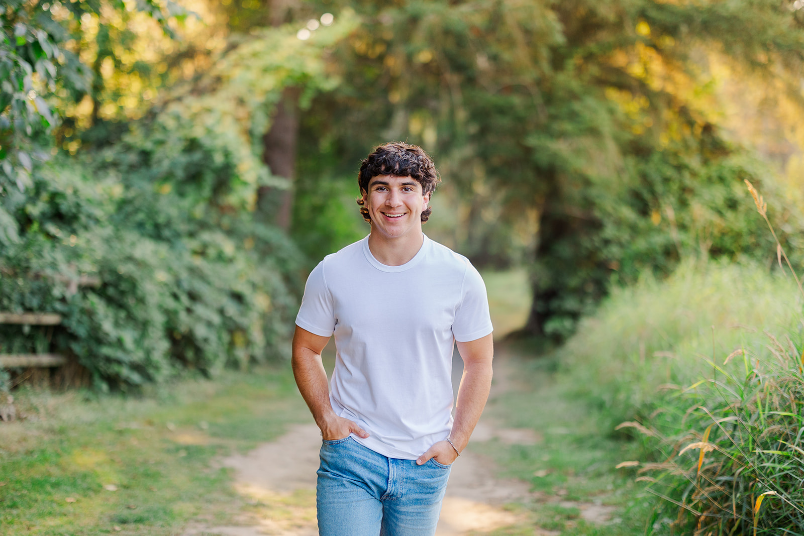 A happy high school senior walks in jeans and a white shirt with hands in pockets on a trail at sunset after visiting the Jewish Community Center Mercer Island