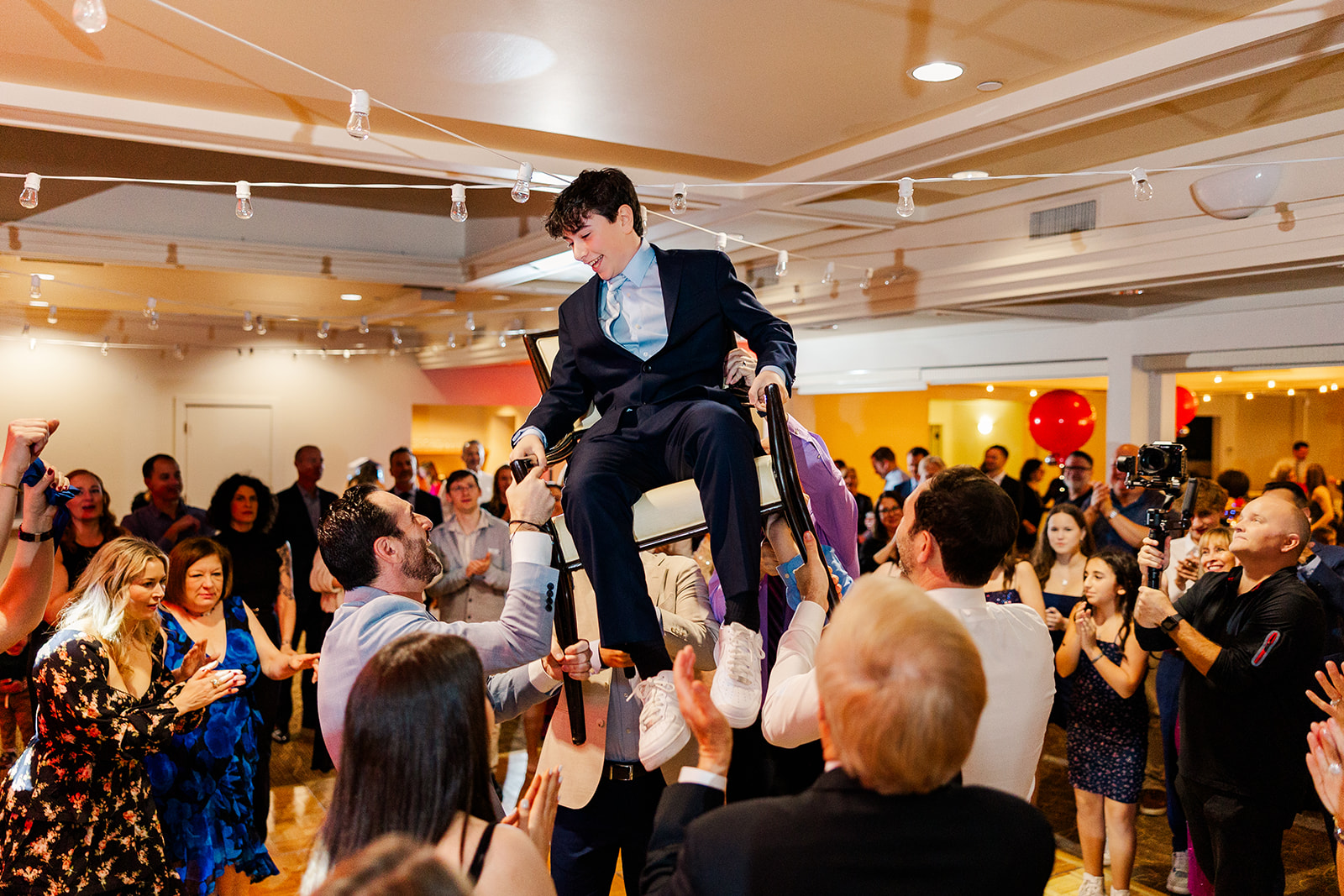 A teen in a suit sits in a chair being lifted on the dance floor at one of the event venues in Bellevue, WA