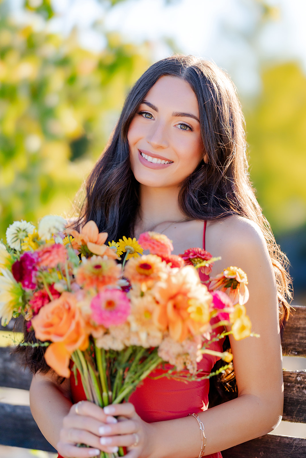 A high school graduate in a red dress sits on a wooden bench holding a bouquet of colorful flowers