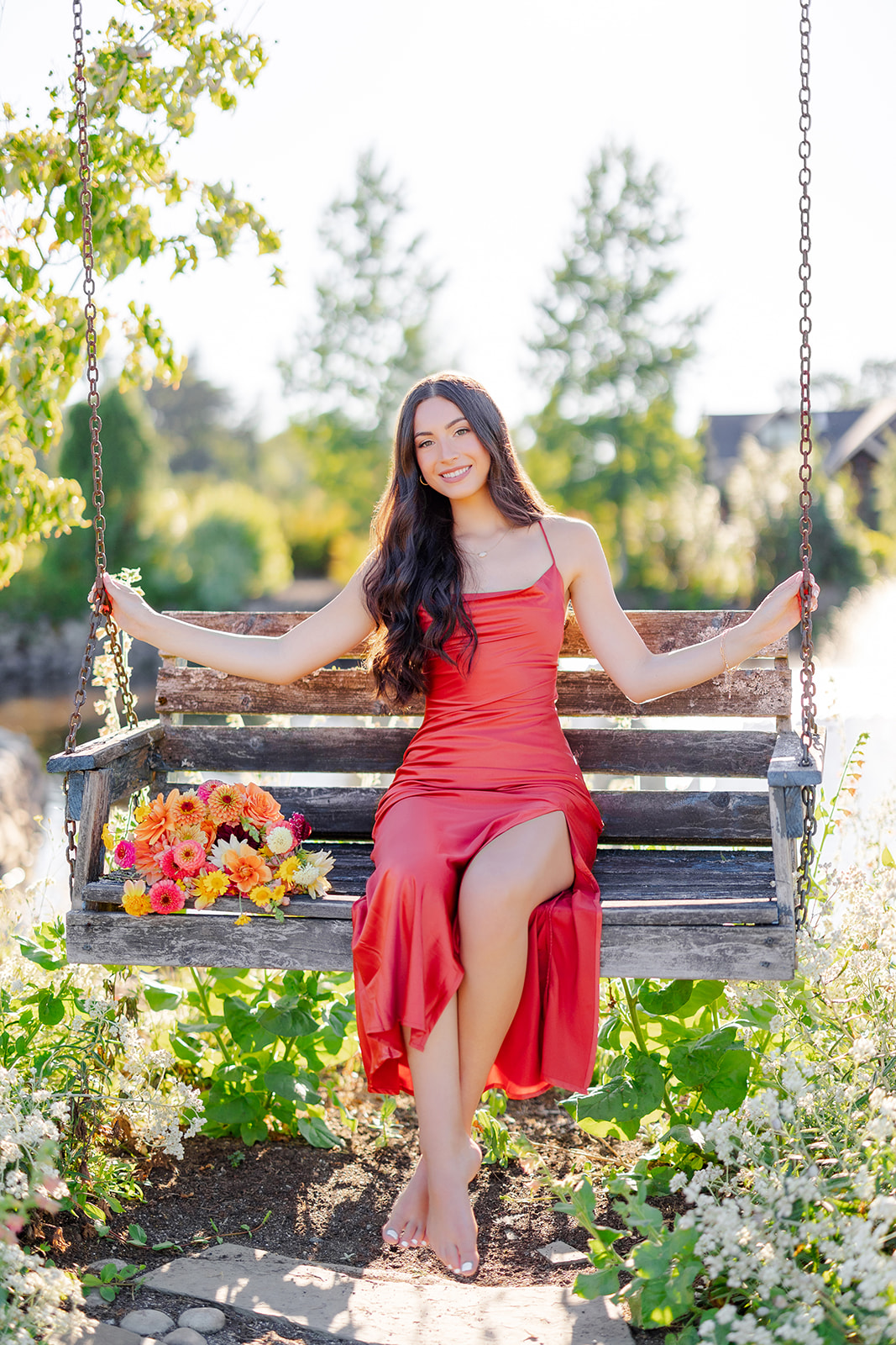 A high school senior in a red dress sits in a garden swing bench with a bouquet from a florist in Mercer Island, WA