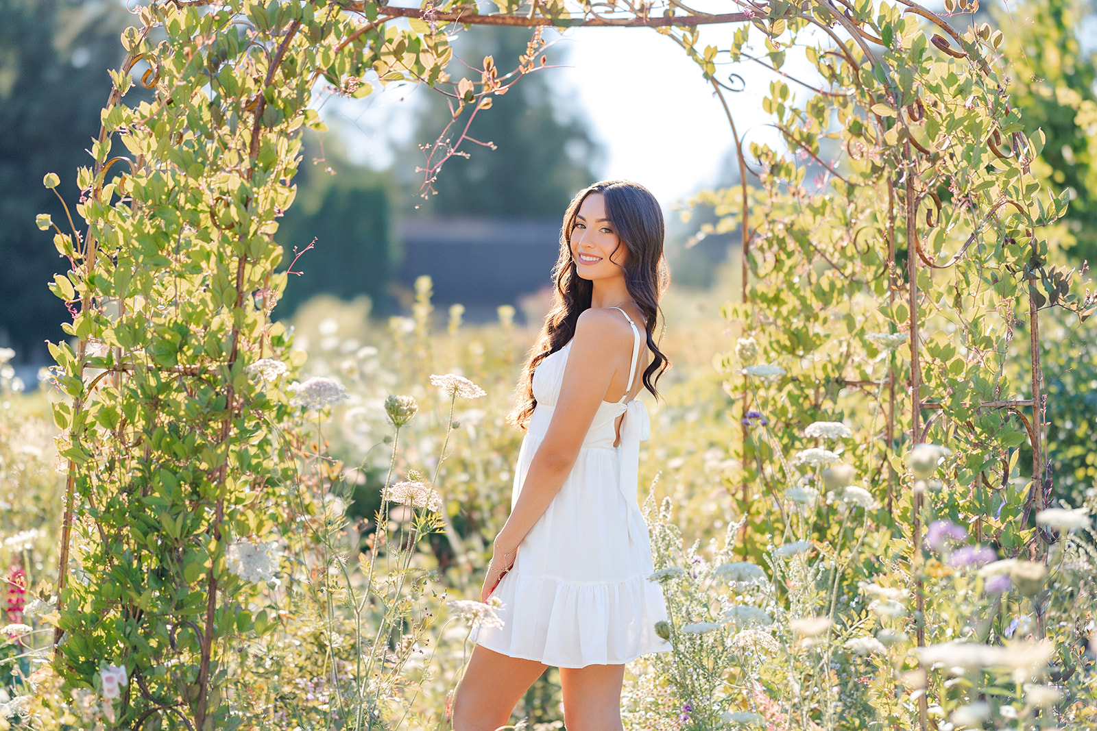 A high school senior stands under a vine covered arch in a white dress smiling after visiting a florist in Mercer Island, WA