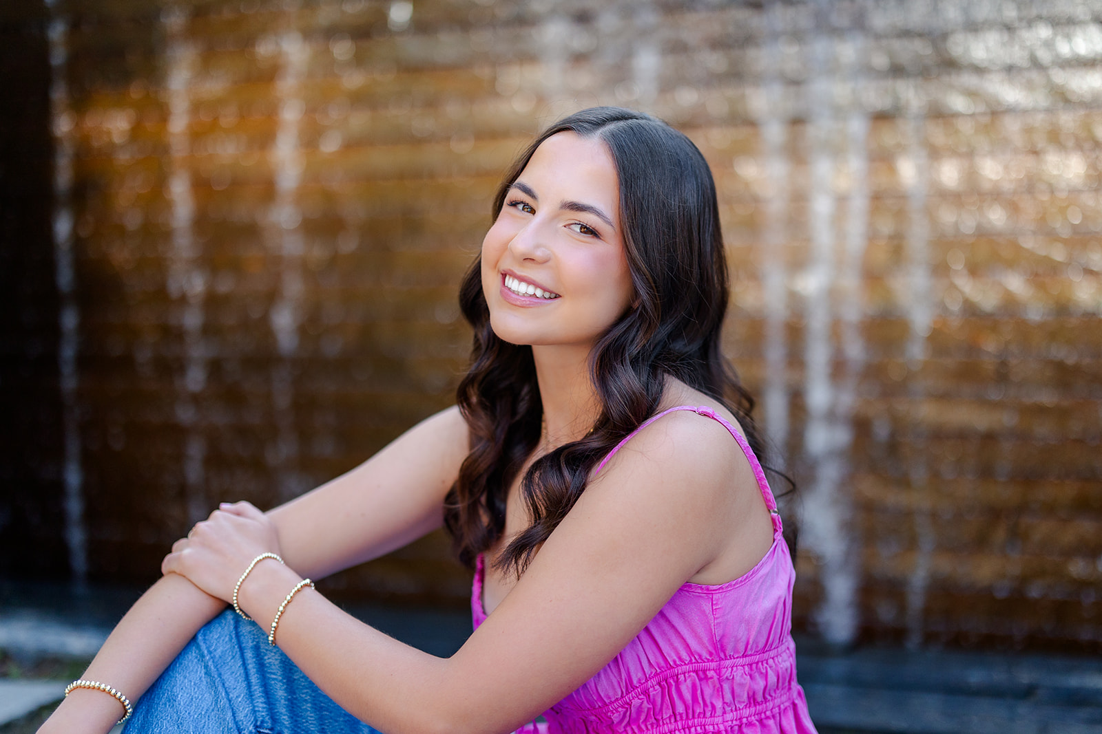 A happy high school senior smiles while sitting in an alley in a pink top