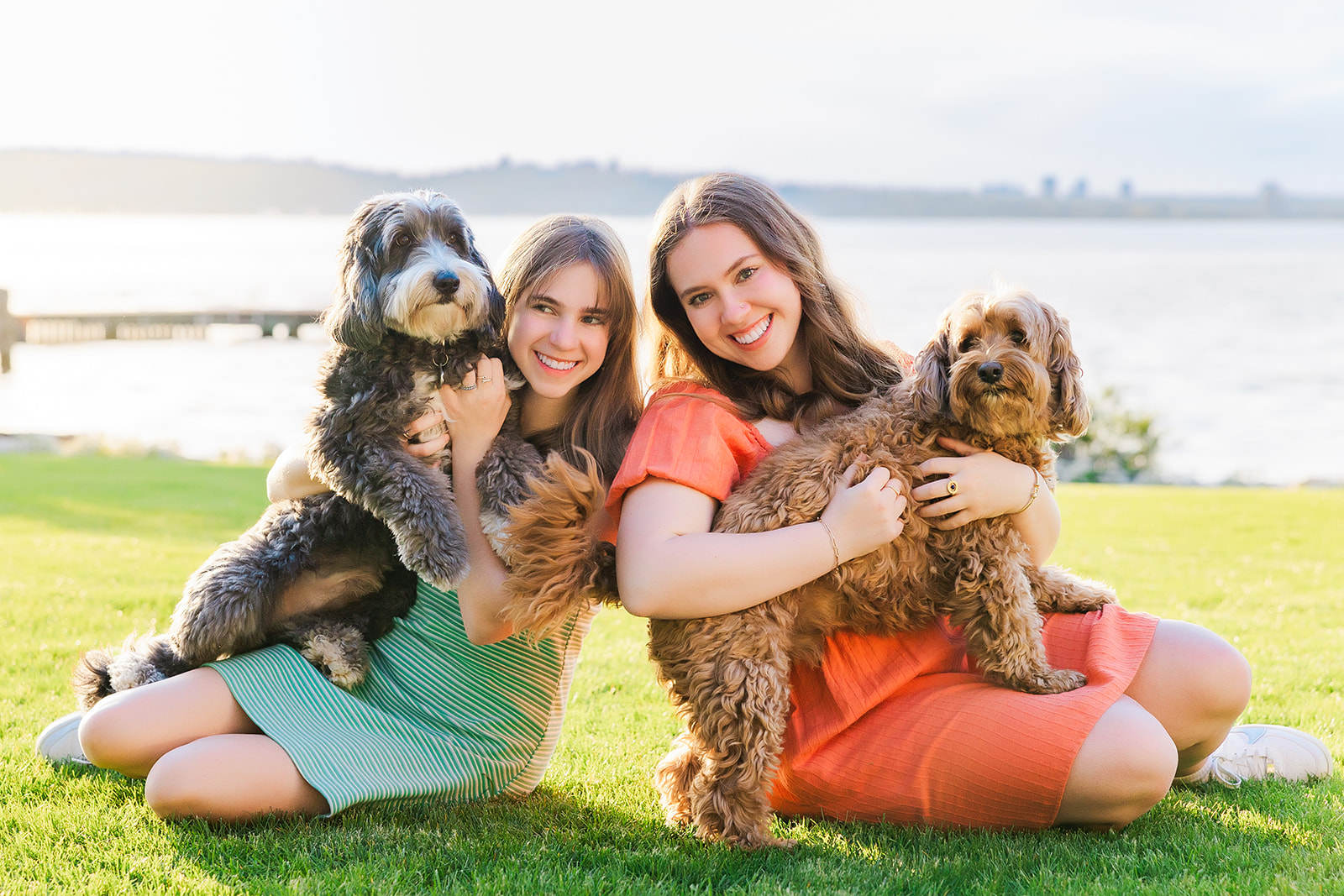 Two teen sisters sit in a waterfront lawn hugging their two dogs after using makeup artists in Kirkland