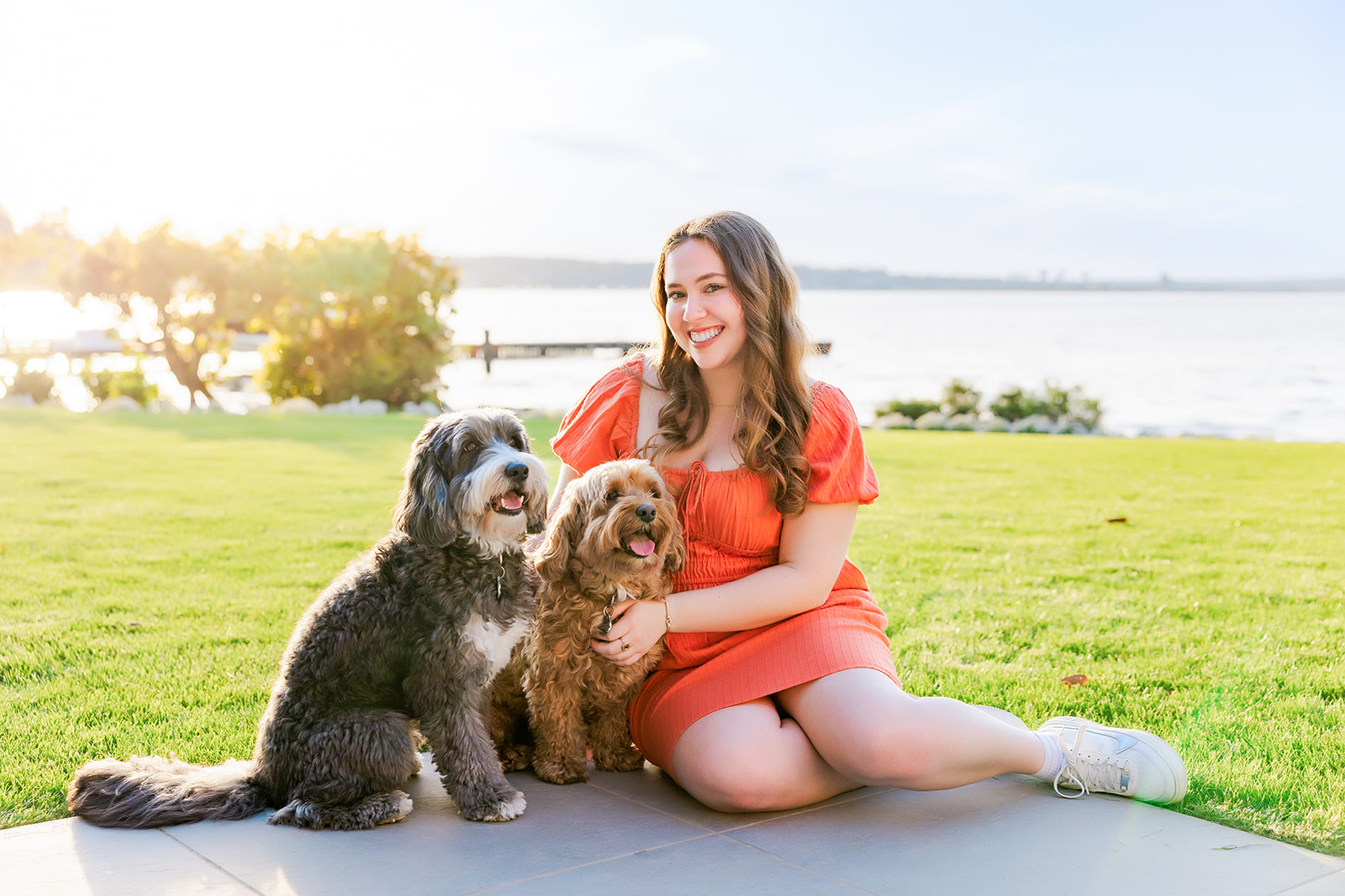 A woman in a pink dress sits on a patio by the water with two dogs after using makeup artists in Kirkland