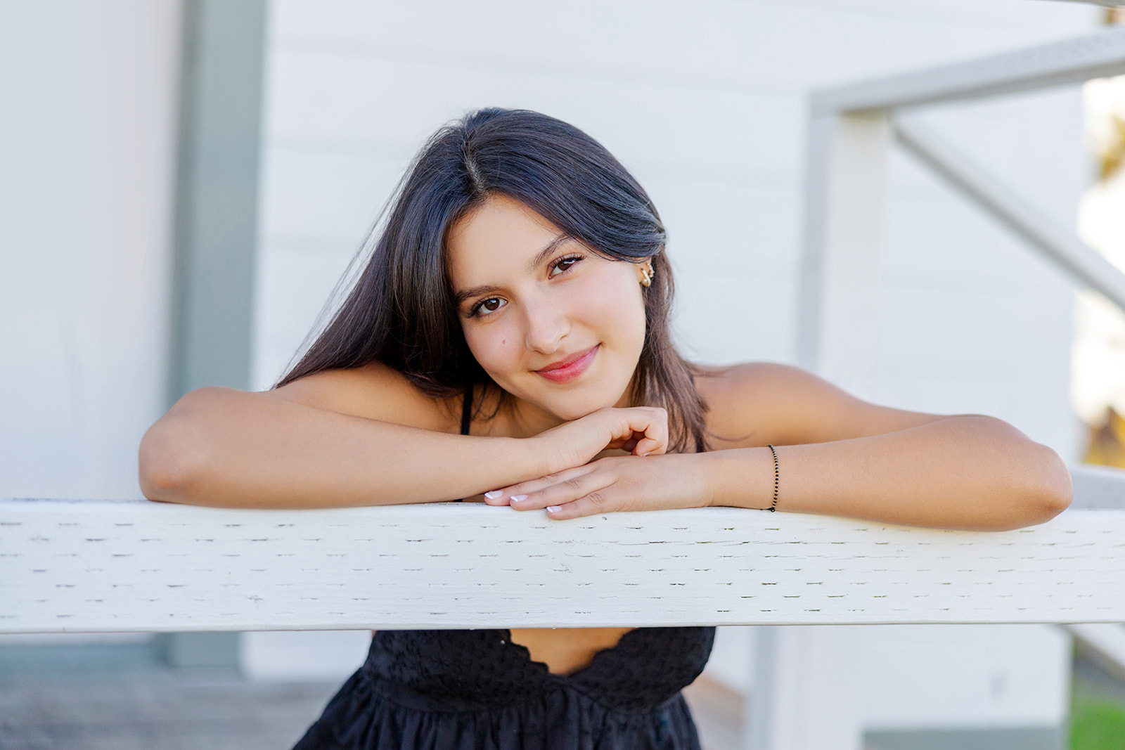 A high school senior leans on a white wooden railing in a black dress