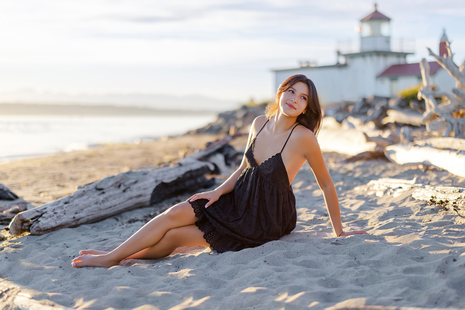 A high school senior in a black dress sits back in the sand on a beach at sunset after visiting makeup artists in Mercer Island