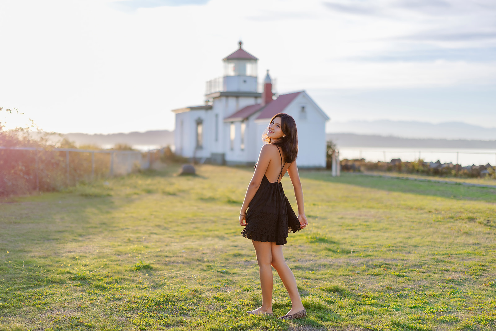 A high school senior smiles over her shoulder while walking in a field of grass by a lighthouse after using makeup artists in Mercer Island