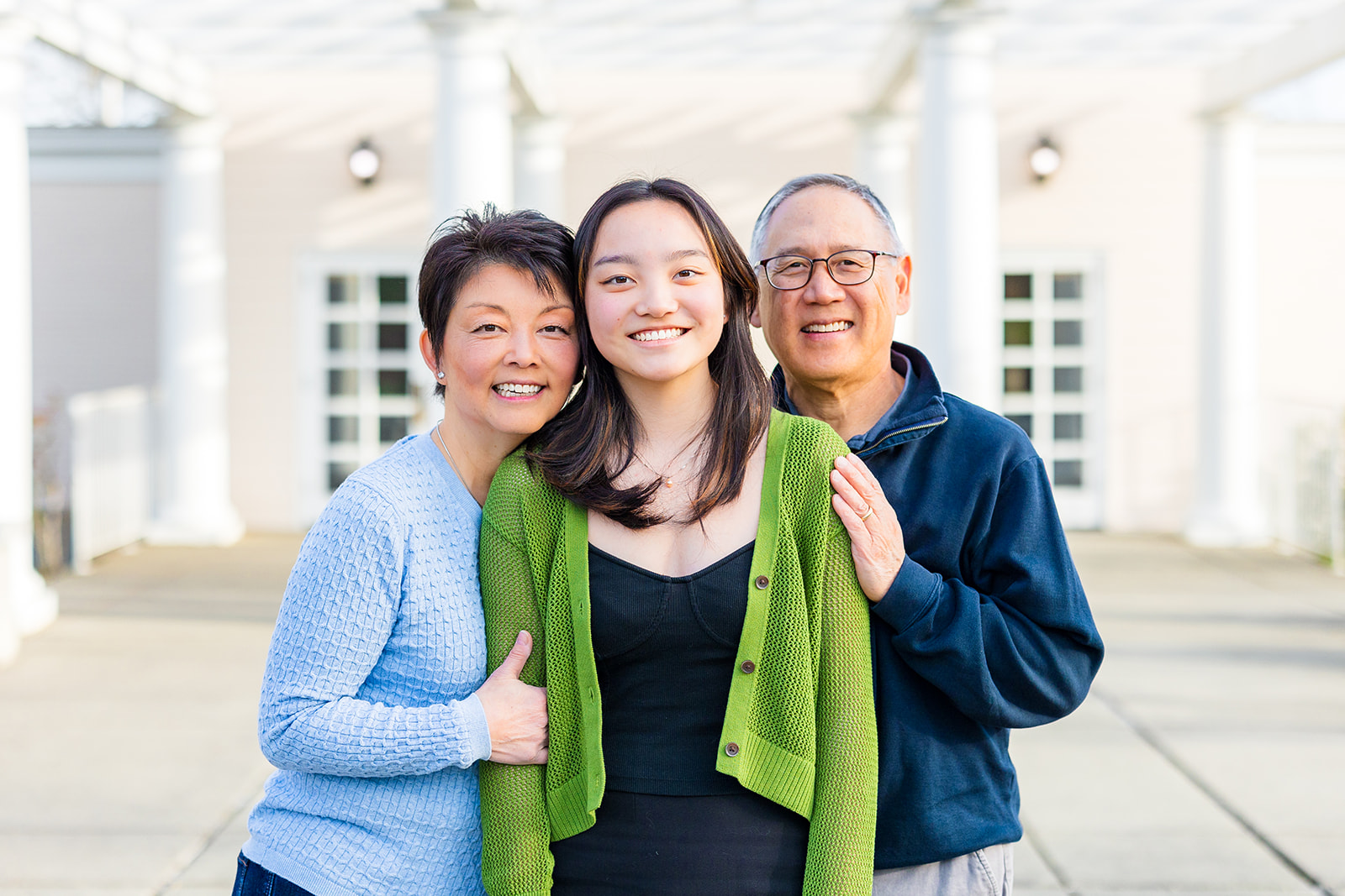 Happy mom and dad lean on their smiling high school senior daughter in a garden under a pergola