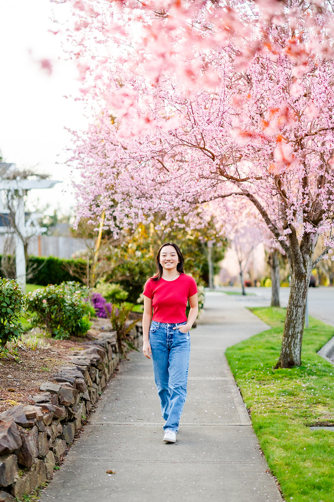 A teenage girl in a red top and jeans walks on a sidewalk under pink blooming trees after using makeup artists in Renton, WA