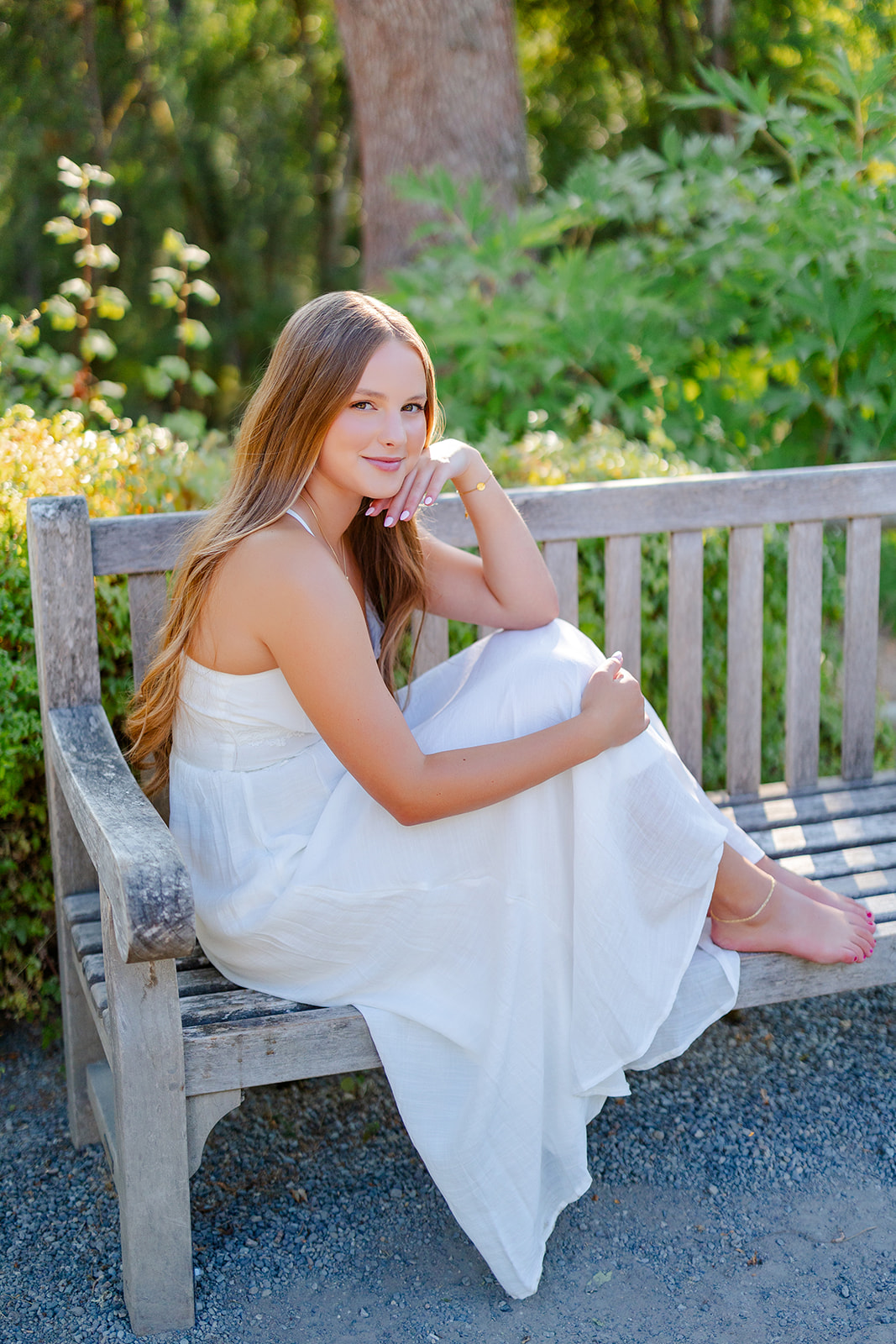 A graduate in a white dress sits on a wood garden bench at sunset