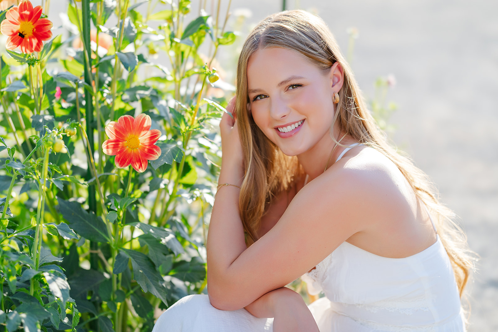 A happy high school senior in a white dress kneels in the sun by son colorful flowers after visiting a nail salon in Sammamish