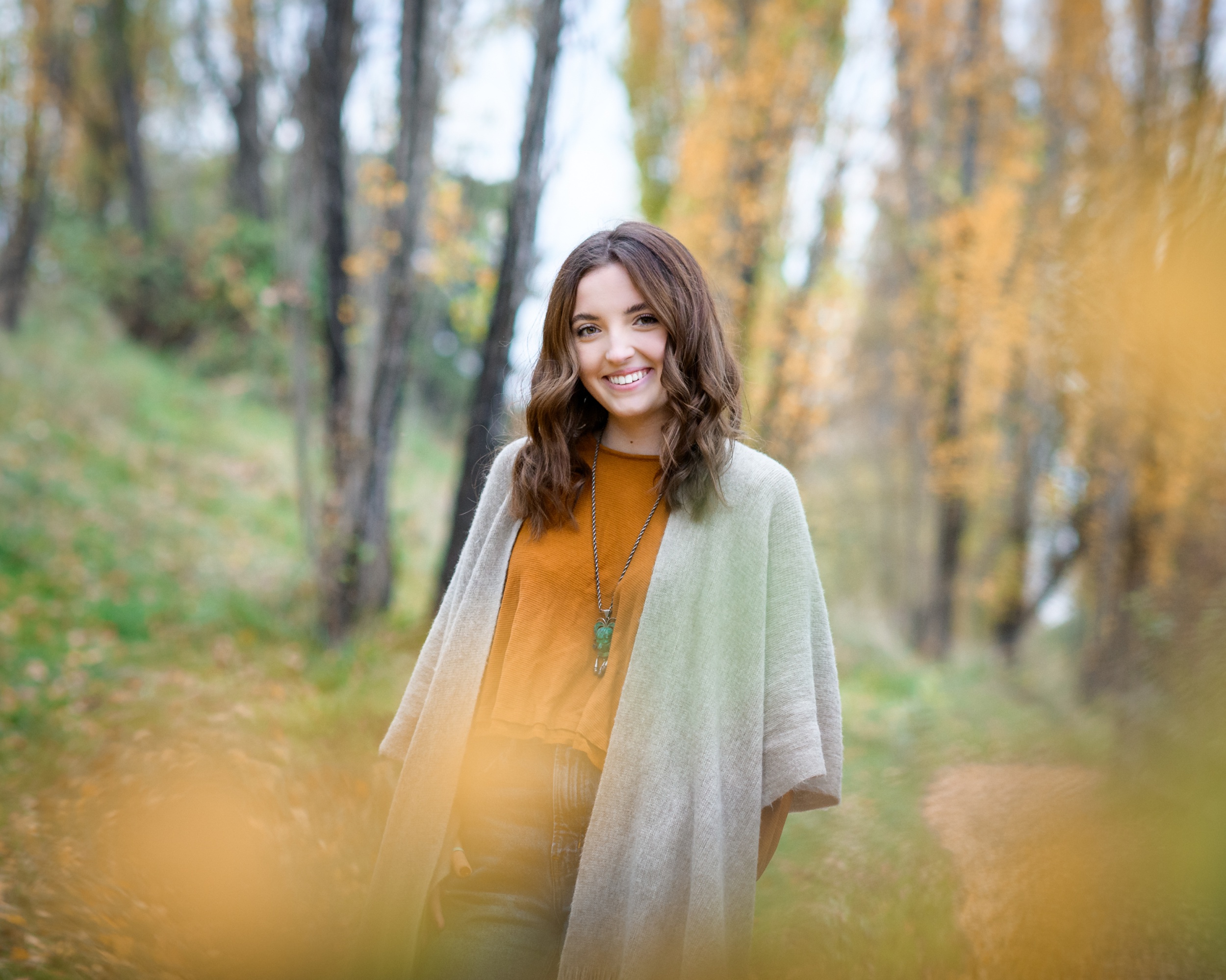 A teenage girl in a grey sweater and orange blouse walks in a forest trail in fall smiling