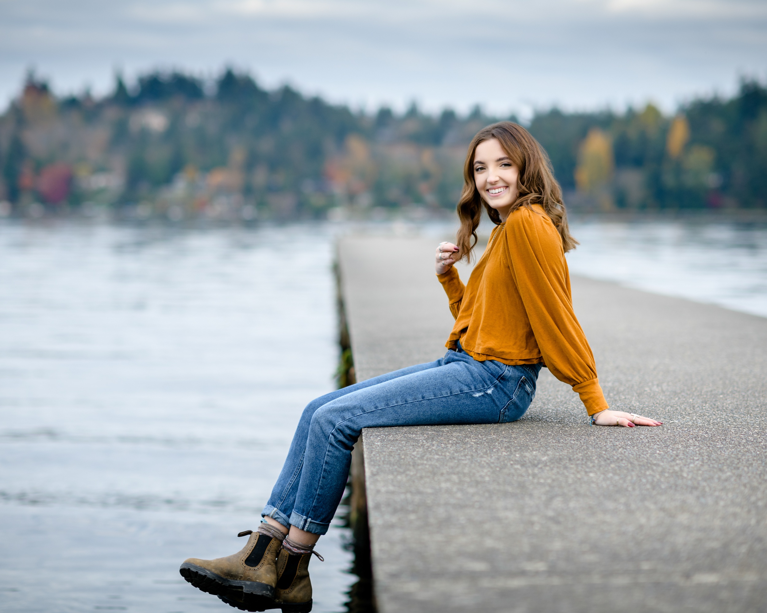 A smiling high school senior sits on a concrete dock in an orange blouse and jeans