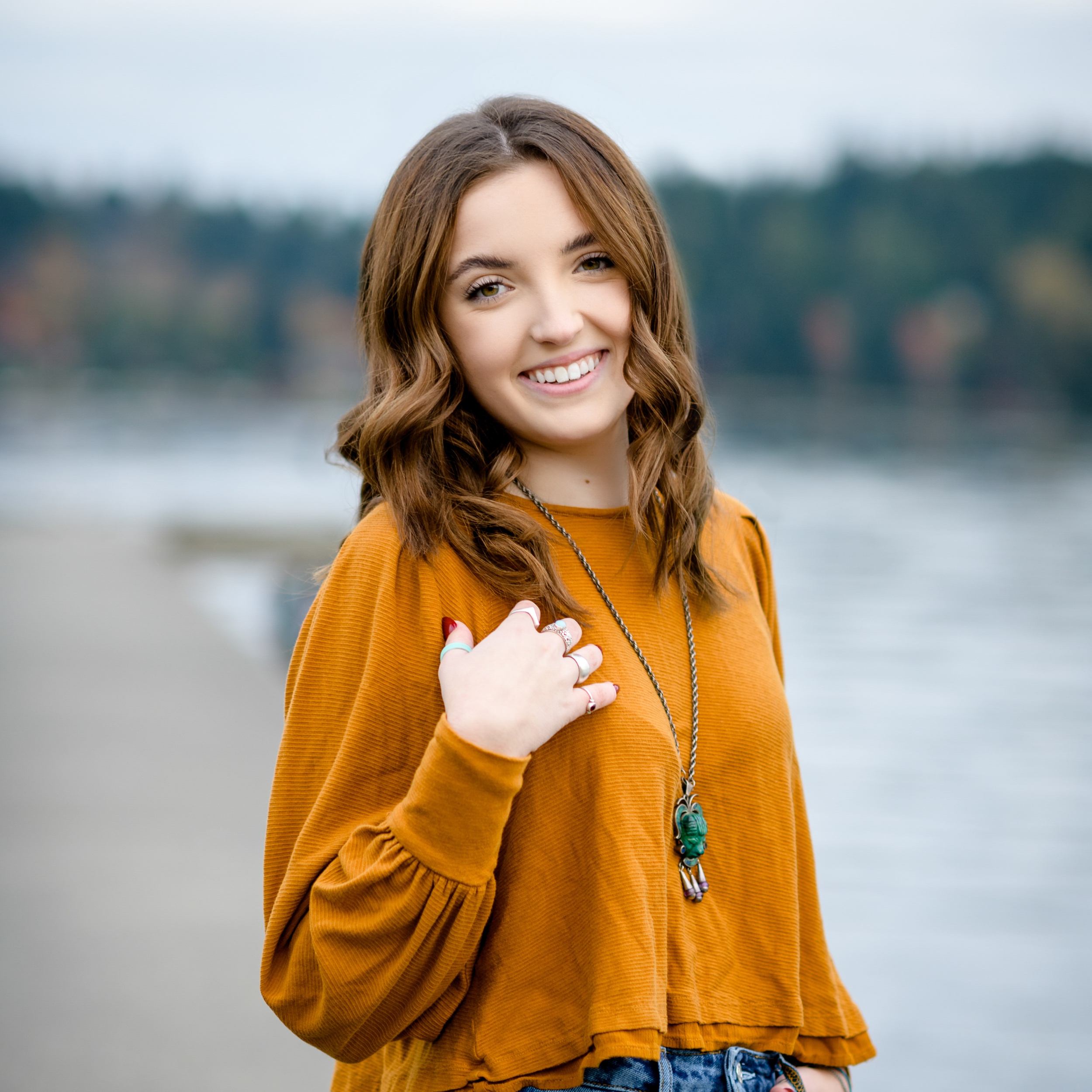 A happy Bellevue Christian School senior smiles in an orange blouse while walking by the water