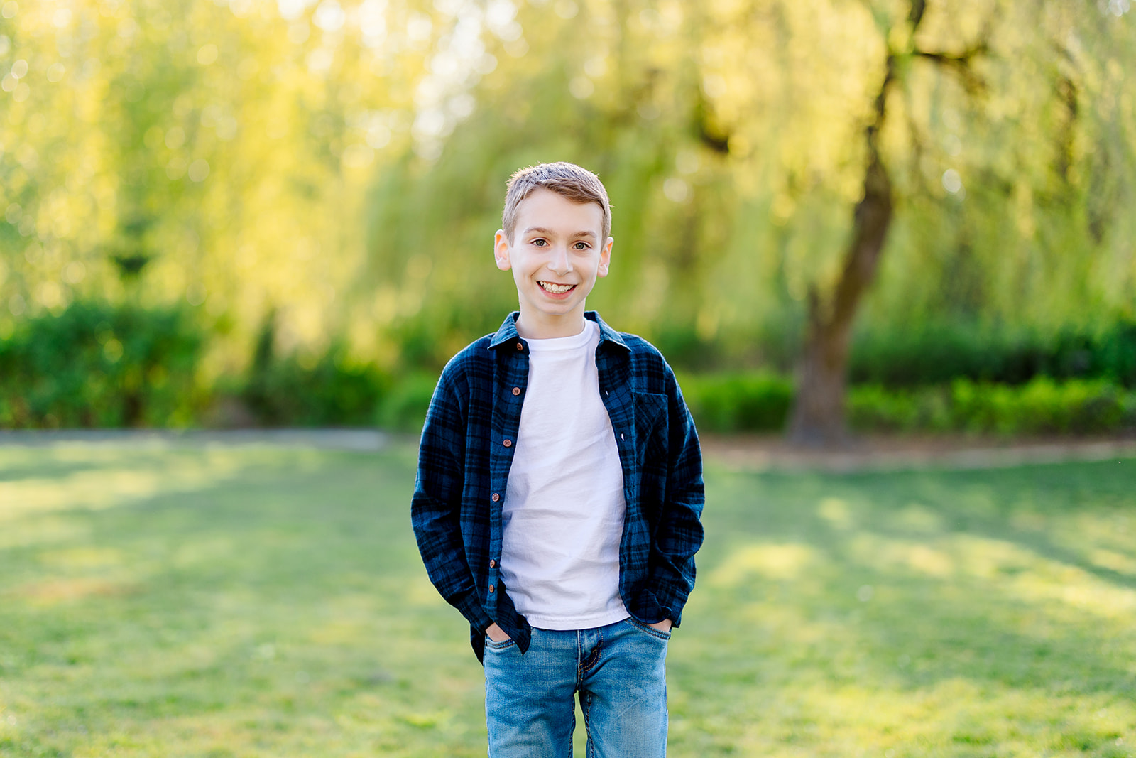 A happy young boy stands in a park lawn at sunset with hands in his pockets
