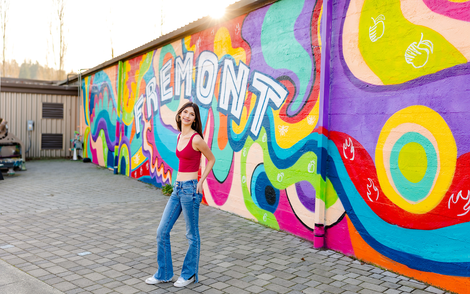A teenage girl in a red top and jeans stands with hands in her back pockets in front of a large mural at sunset