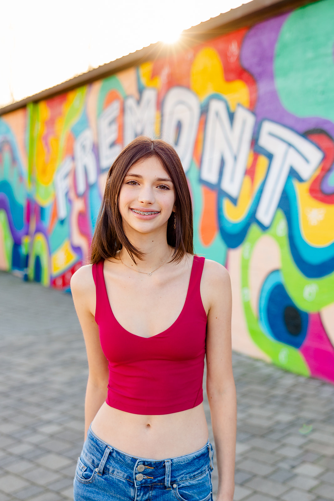 A high school senior in a red top and jeans stands smiling in front of a mural at sunset after finding Catering in Sammamish