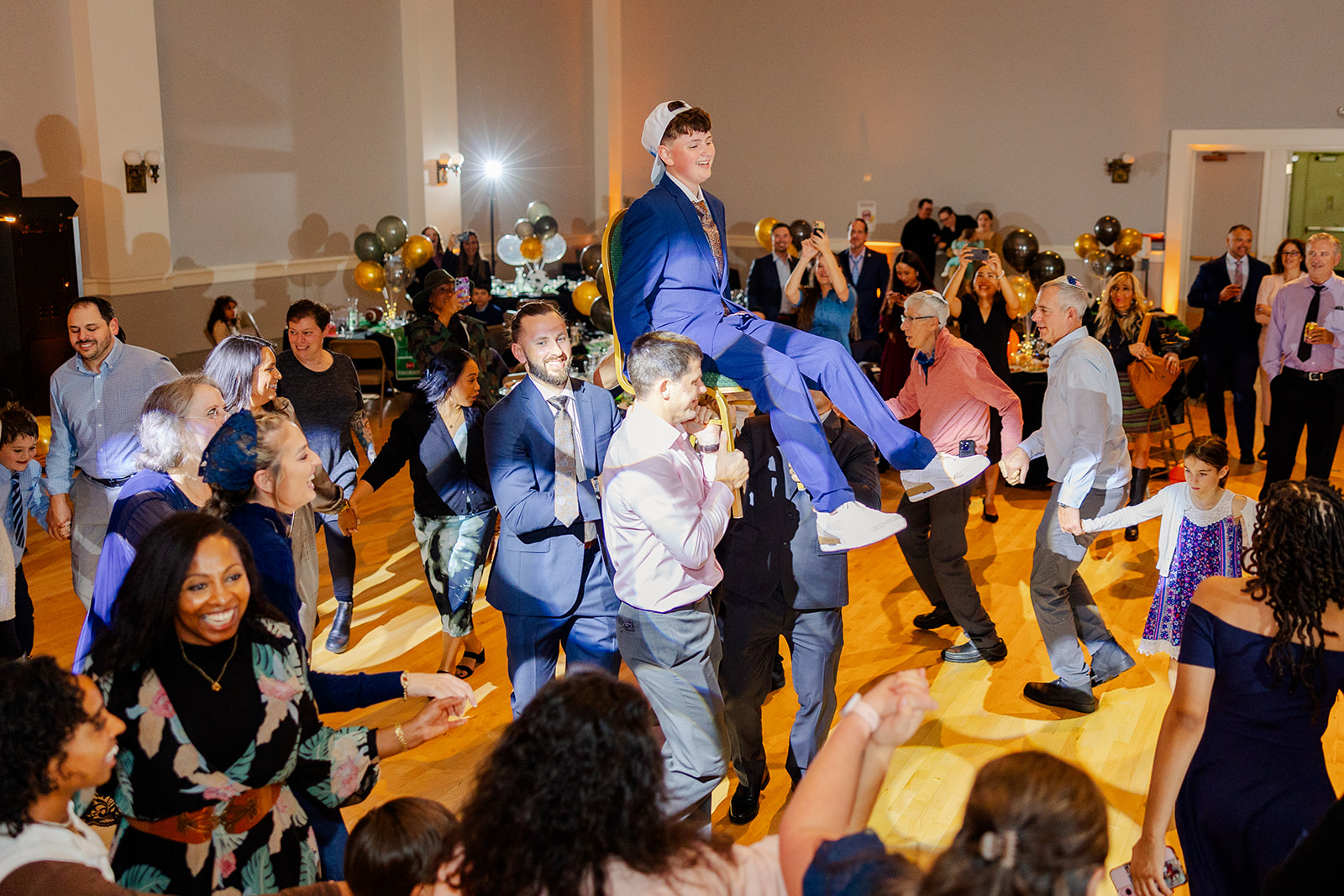 A teen in a blue suit sits on a chair held up by adults on the dance floor with everyone around them