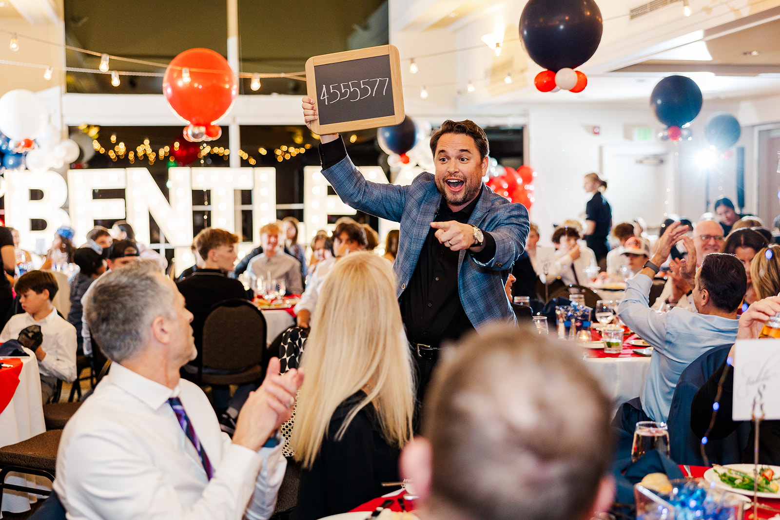 A man in a blue plaid suit holds up a chalkboard in a Bar Mitzvah reception