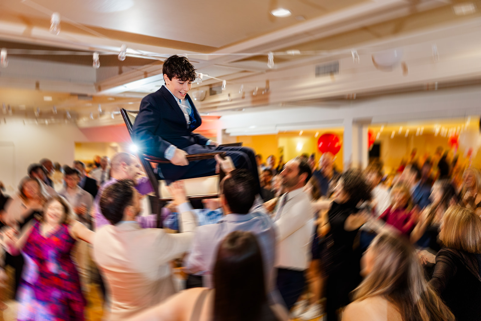 A boy sits in the chair while being lifted on the dance floor in a blue suit