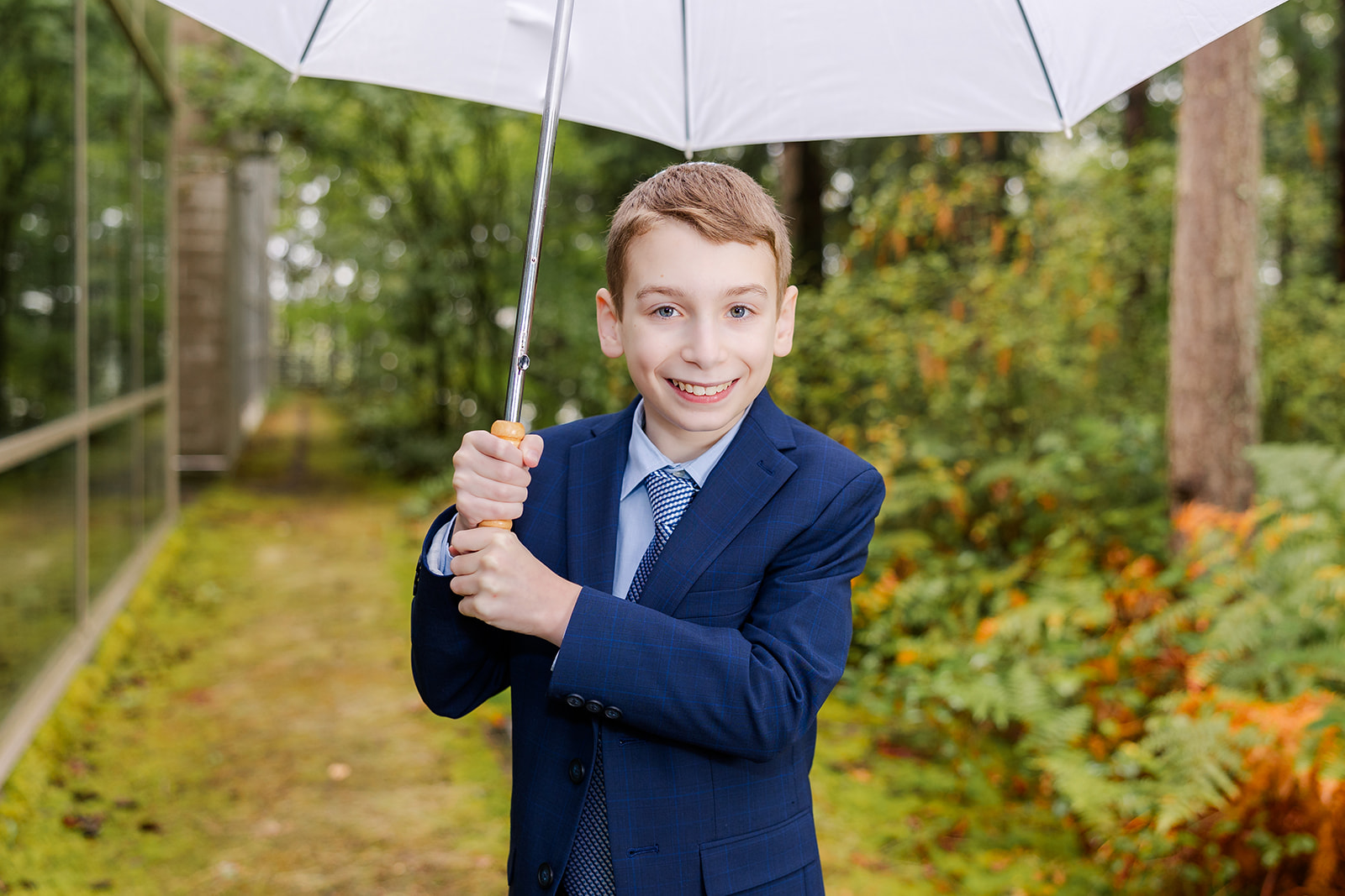 A young boy in a blue suit holds an umbrella while smiling in a forest behind a glass building after meeting event planners in Seattle