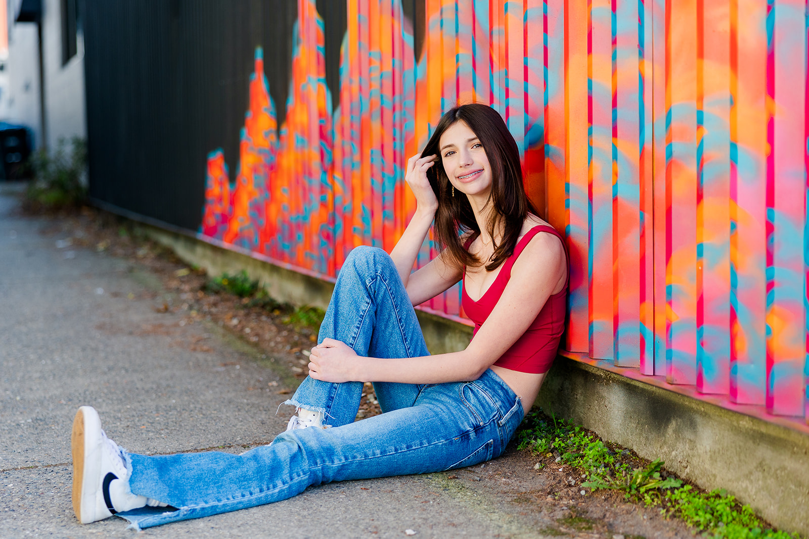 A high school senior in jeans and red top sits against a mural wall in an alley