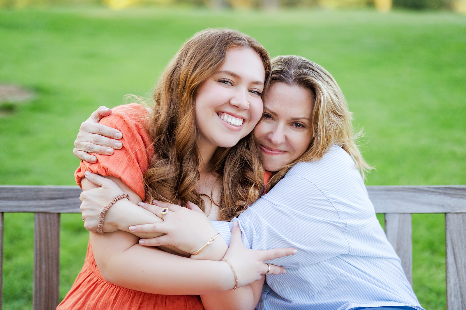 A proud mom hugs her graduating teenager in a pink dress on a park bench