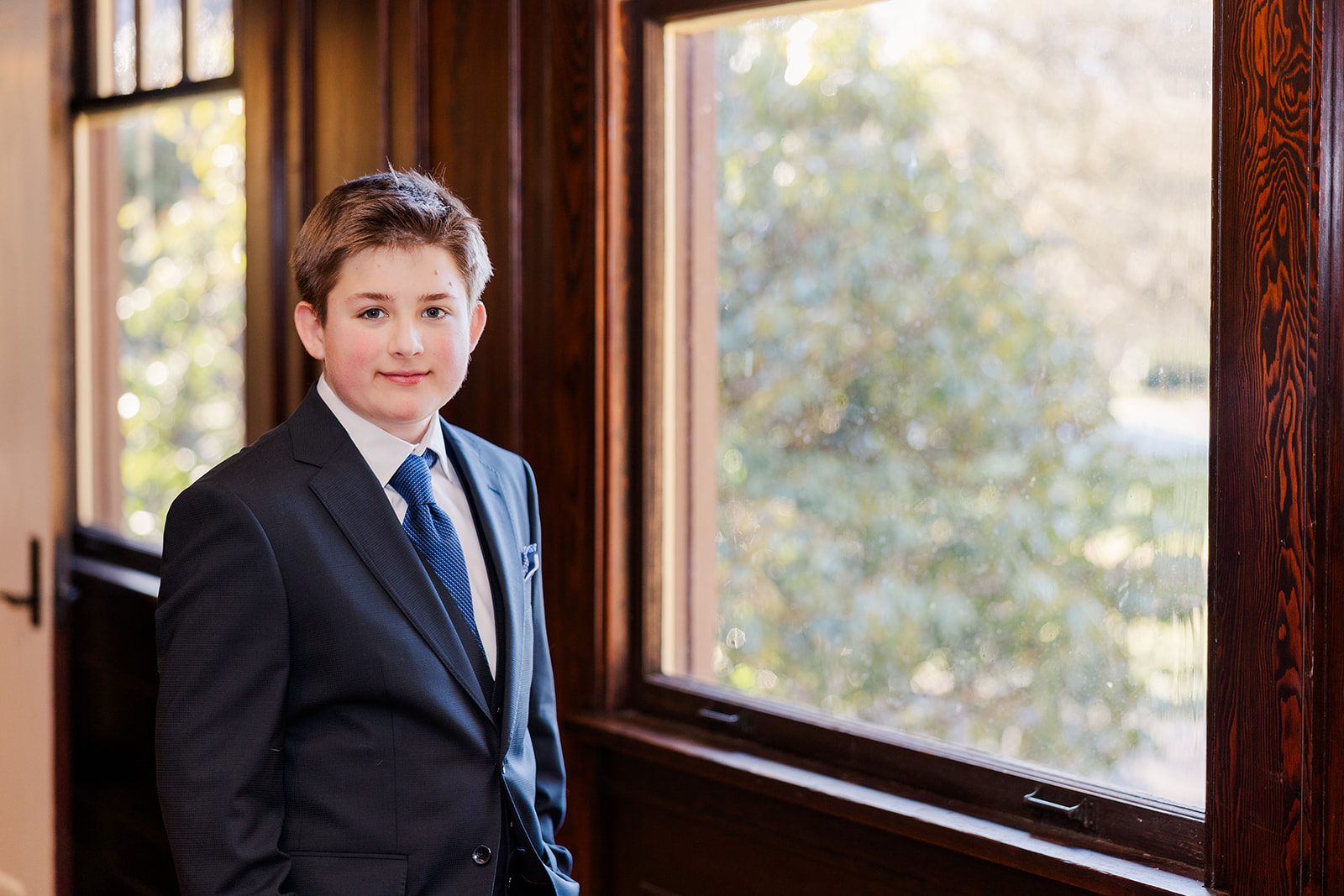 A boy in a blue suit stands in a window smiling with hands in his pockets after finding a bakery in Kirkland, WA