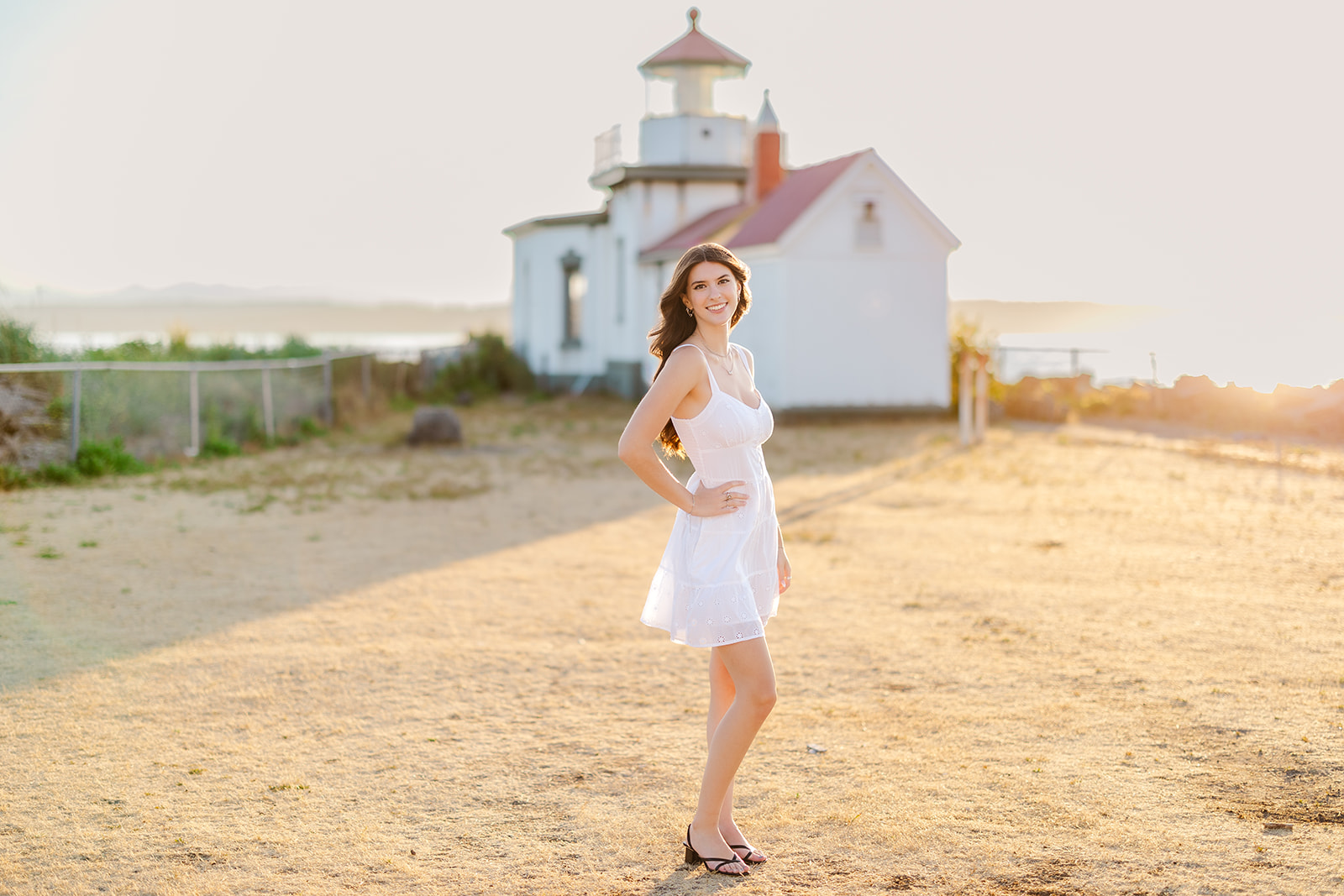 A high school grad in a white dress stands in a dirt lot of a lighthouse at sunset smiling