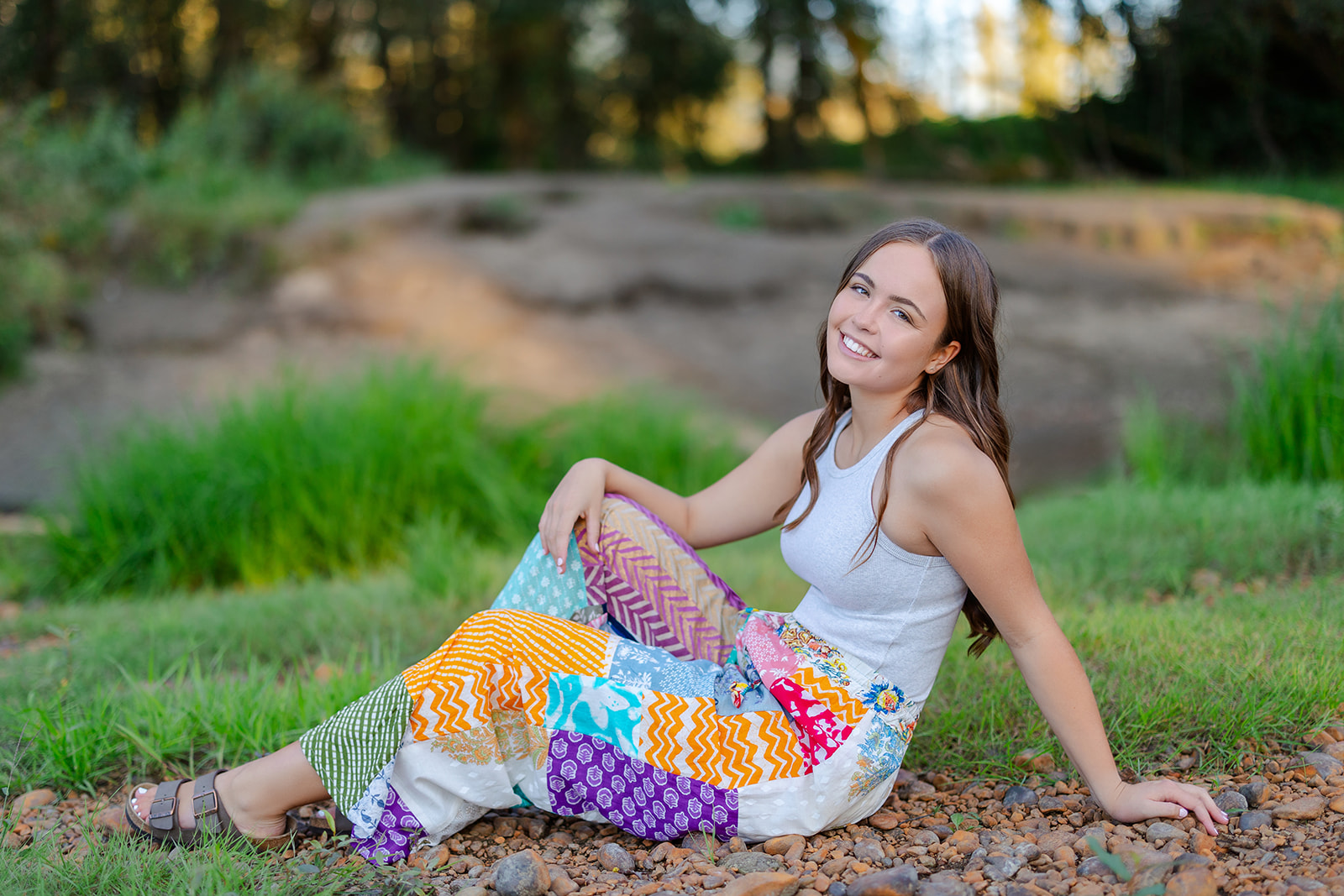 A high school senior in a colorful skirt sits in a park smiling