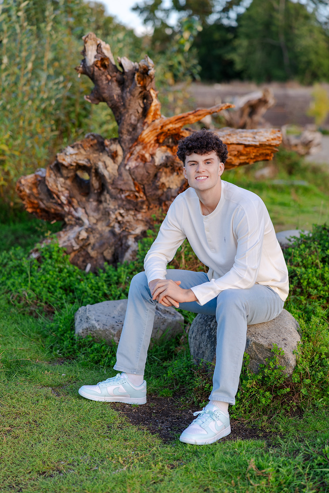 A teenage boy in a white shirt sits on a boulder in a park smiling after attending College in the High School