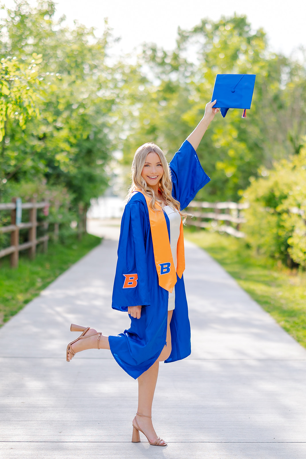 A graduate in her gown and white dress lifts her cap and leg in a park sidewalk after using College in the High School