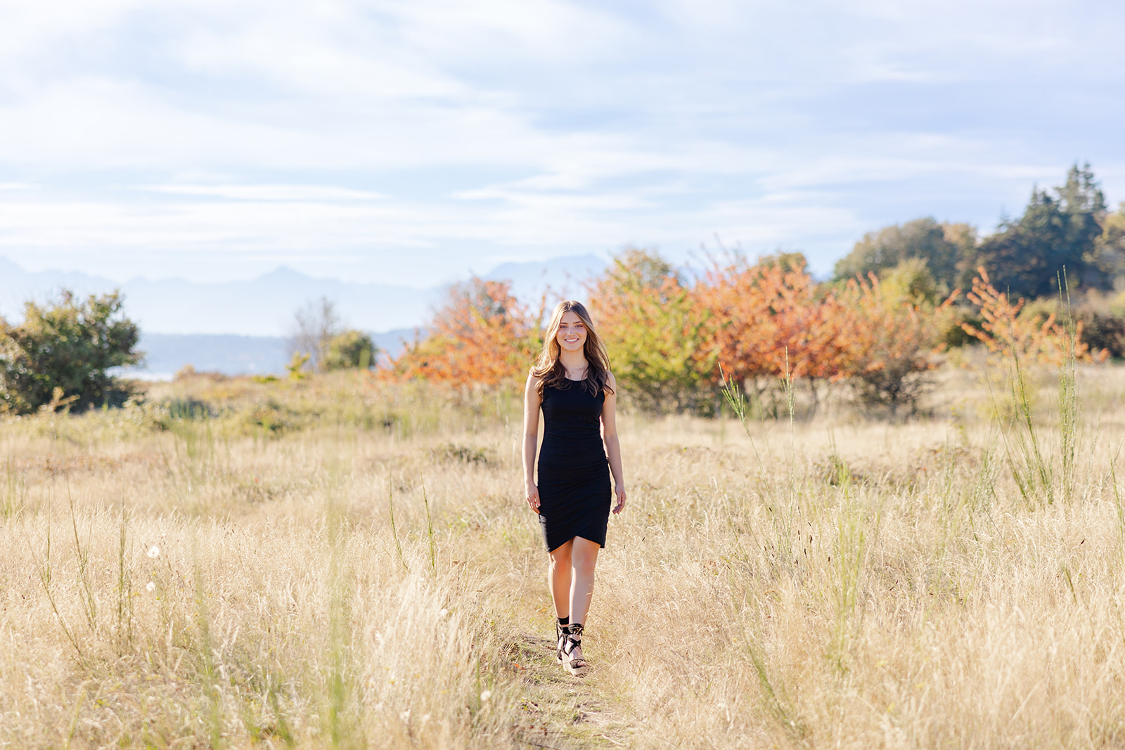 A high school senior in a black dress walks on a trail in a meadow smiling