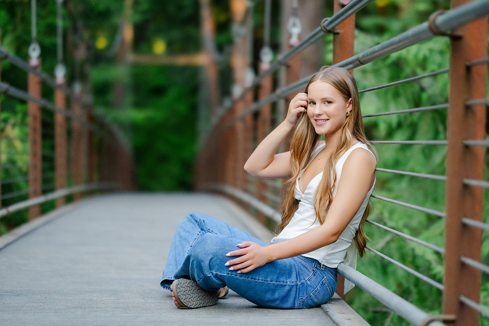 A high school senior in a white top and jeans sits on a pedestrian bridge smiling after visiting colleges in downtown Seattle